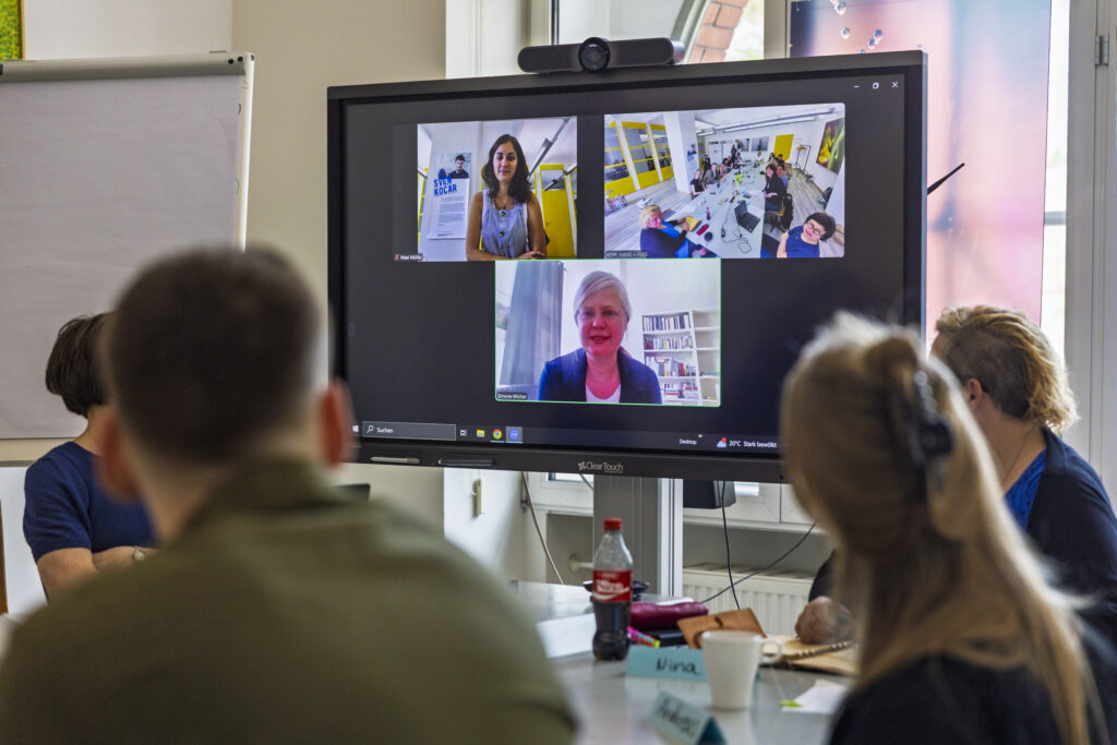 Workshop-Situation: Foto von Personen, die an einem Tisch sitzen und gemeinsam auf einen Screen mit weiteren Online-Teilnehmenden schauen.