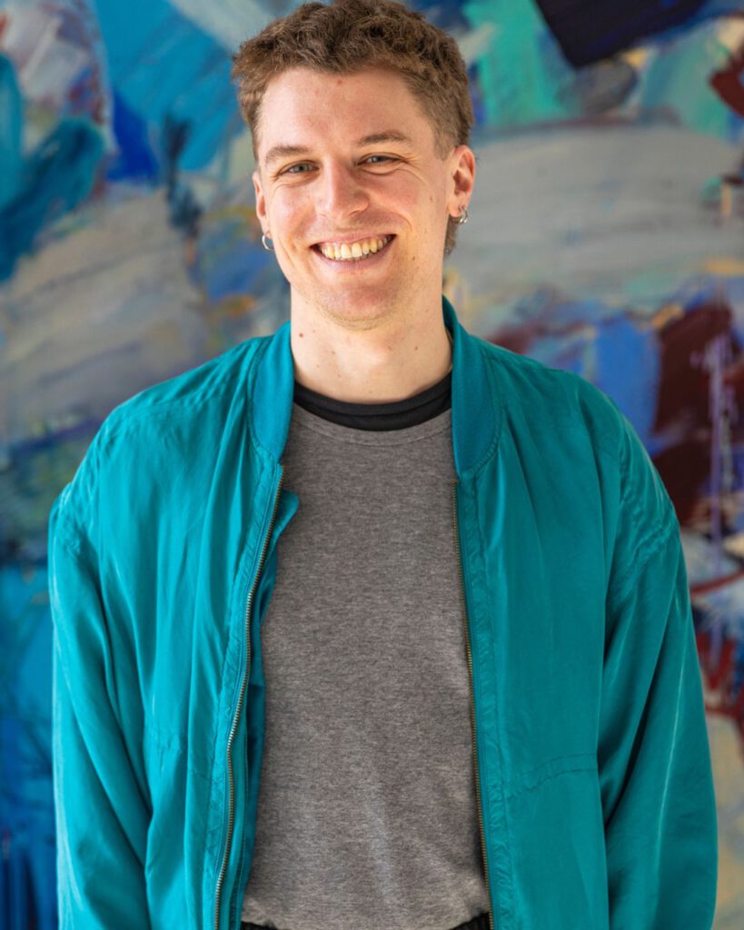 junger weißer Mann mit blonden Haaren und blauer Jacke.